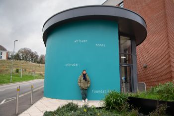 Helen Cammock stands in front of the artwork on an external wall at top of ɫ Student Centre. The text is painted in white and set against a bright teal background, and reads: whisper  tones  vibrate  foundations